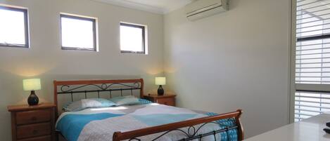 A bedroom with a bed, two bedside tables with lamps, a window with blinds, and an air conditioning unit.