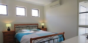 Un dormitorio con una cama, dos mesitas de noche con lámparas, una ventana con persianas y un equipo de aire acondicionado.