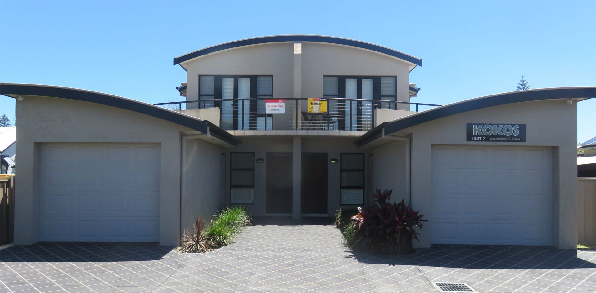 Une maison moderne à deux étages, avec un garage de chaque côté, un balcon à l’étage supérieur et une enseigne indiquant « Location de vacances ».