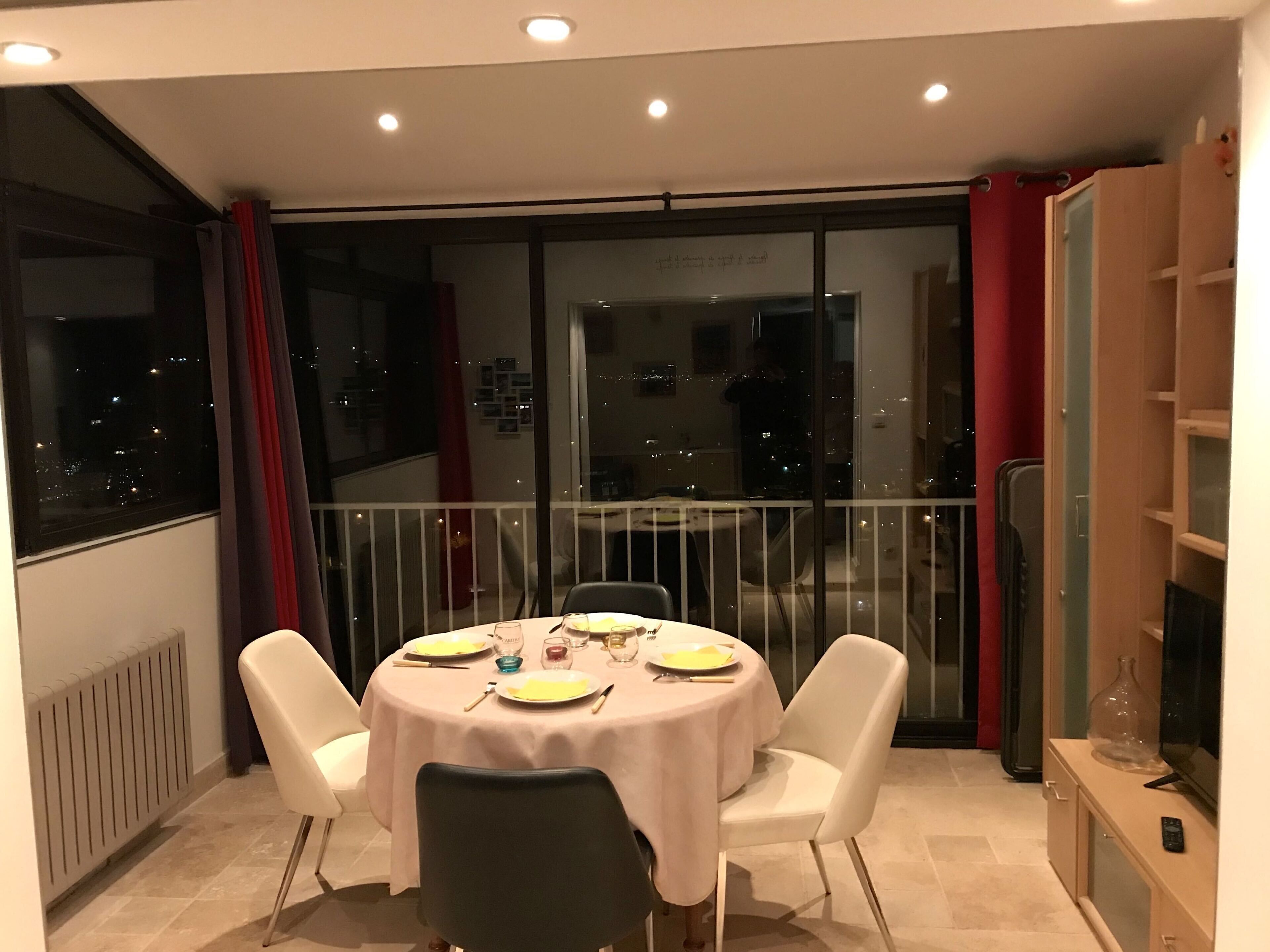 Ein Essbereich mit einem runden Tisch, weißen Stühlen und Blick auf die beleuchtete Stadt durch große Fenster.