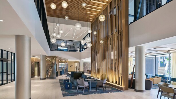 Un lobby espacioso con un diseño moderno, que cuenta con un techo alto con luces esféricas colgantes, una escalera y un área de estar con mesas y sillas.