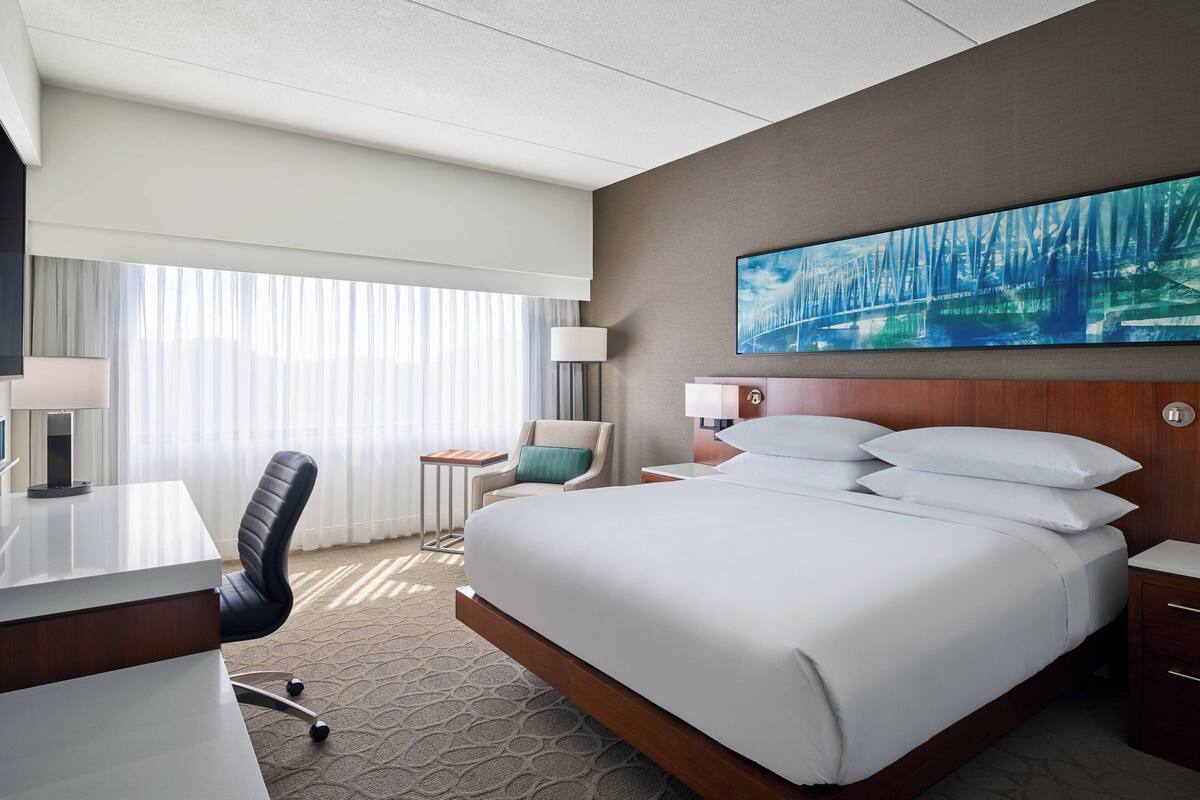 A hotel room with a large bed, a desk, a chair, a small table, and a painting on the wall.