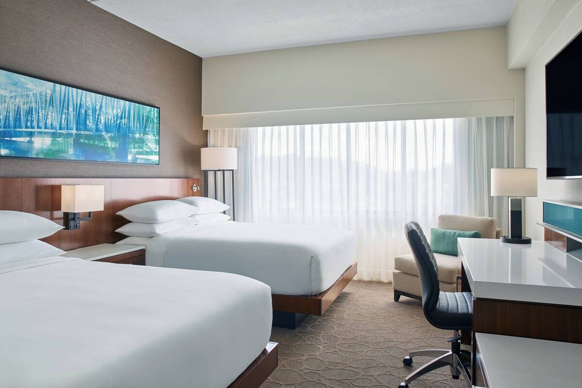 A hotel room with two beds, a desk, a chair, a TV, and a large window with curtains.