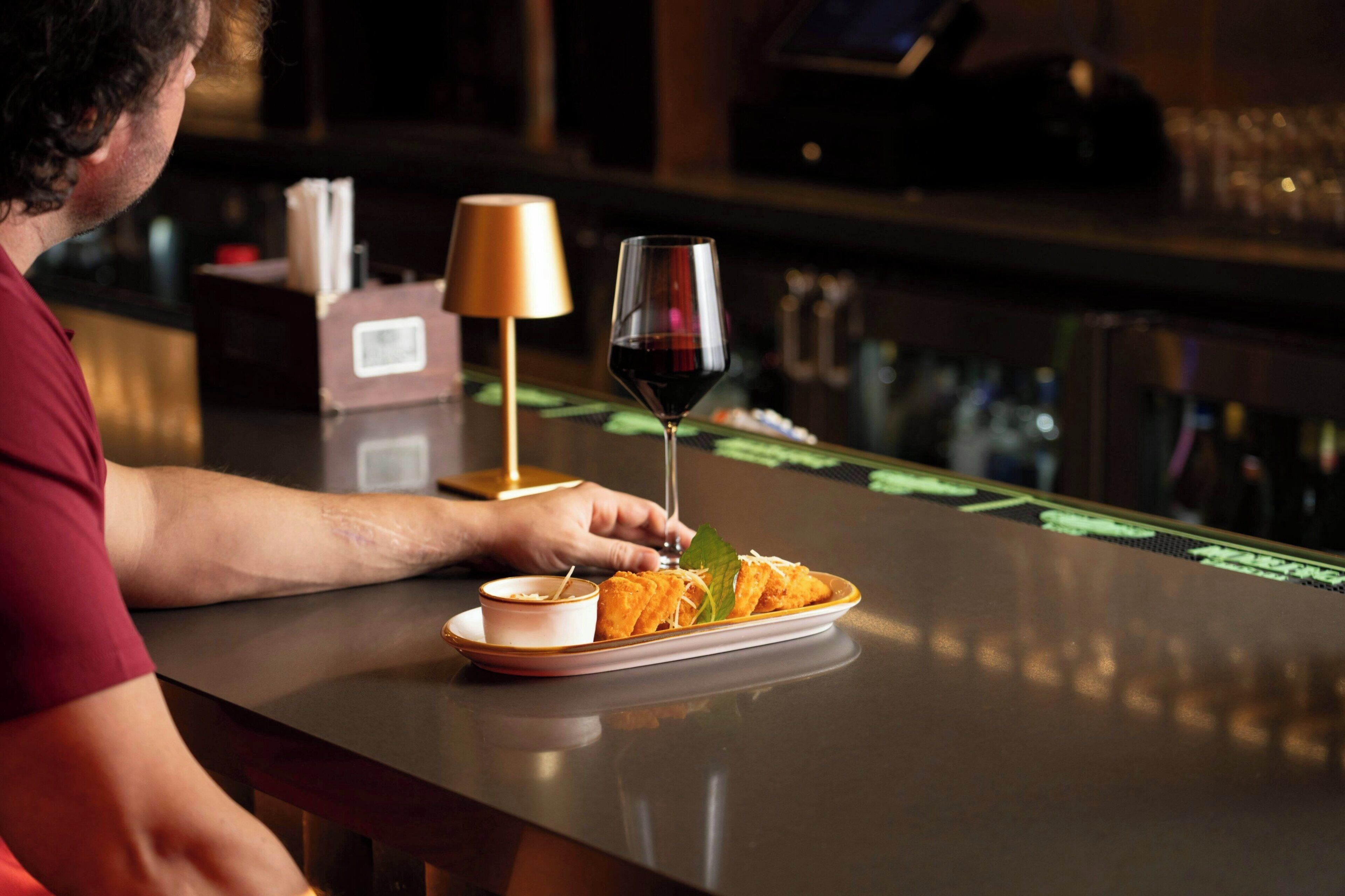 A person seated at a bar with a plate of food, a glass of red wine, and a small bowl of sauce.