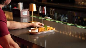 A person seated at a bar with a plate of food, a glass of red wine, and a small bowl of sauce.