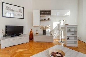 A modern living room with a flat-screen TV, a light wood cabinet, a white shelving unit, and a herringbone wood floor.