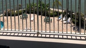 Een balkon met uitzicht op kalm water, palmbomen en een zandstrand met ligbedden.