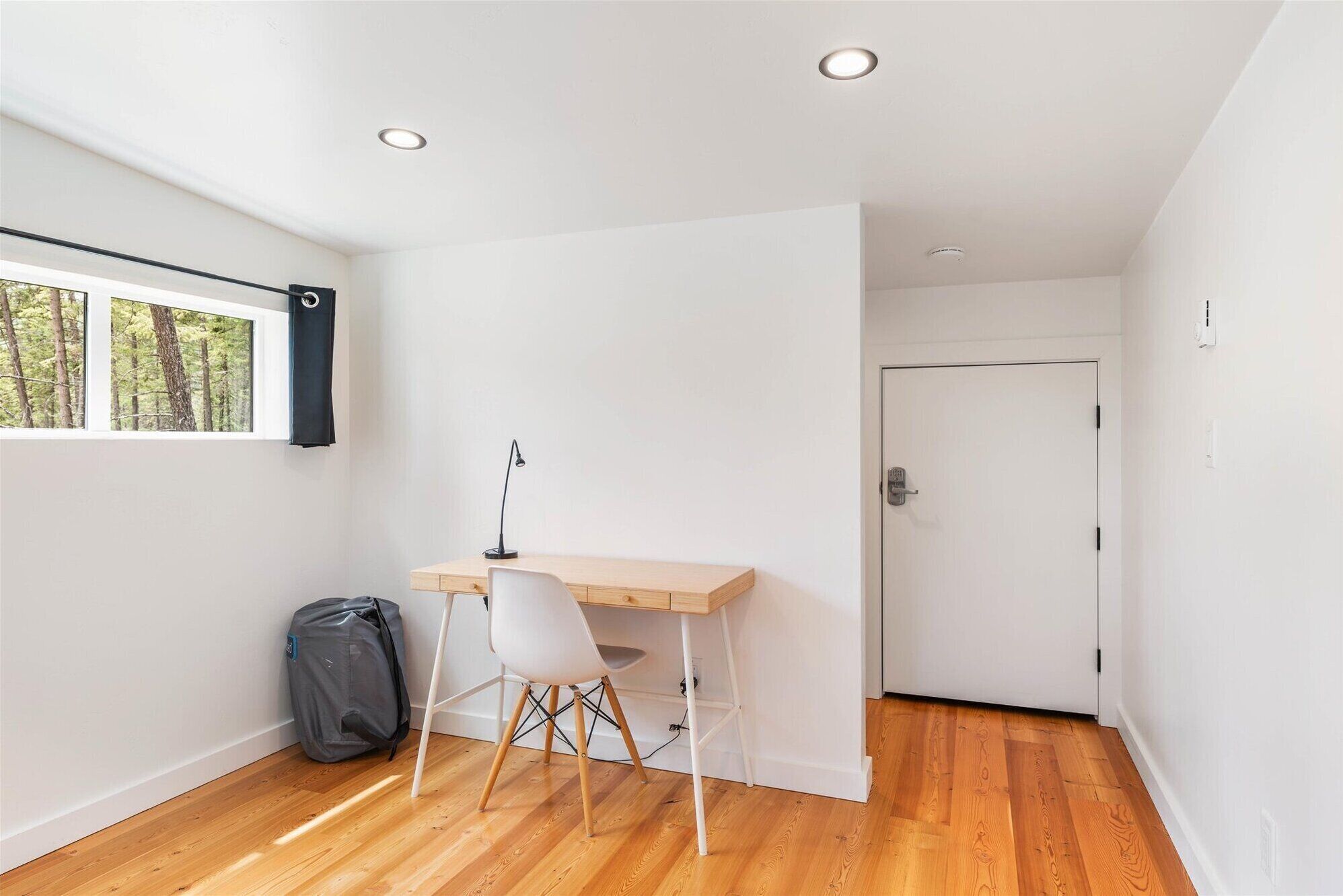 Una habitación pequeña y luminosa con un escritorio de madera, una silla blanca y una ventana con vistas a árboles.