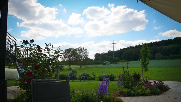 A view from a room with a glass table reflecting the sky and clouds. Outside, there's a green field, a tree, and a cross on a pole. - Vacation apartment Blumenwiese - Preuschens Hof (Egloffstein)
