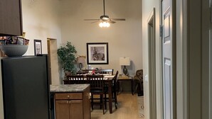 Une cuisine avec des meubles de rangement en bois, une table à manger, des chaises, un ventilateur de plafond et un réfrigérateur.