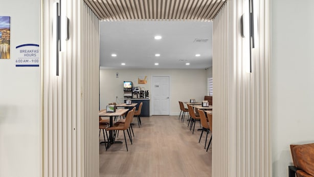 A hotel dining area with tables and chairs, a kitchen in the background, and a sign indicating breakfast hours. - Baymont by Wyndham Orlando Downtown (Orlando)