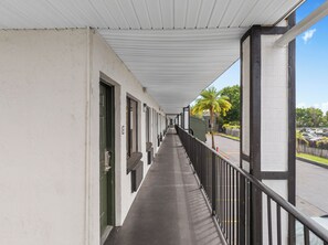 A corridor with a covered walkway, doors, and a view of a parking lot and palm trees.