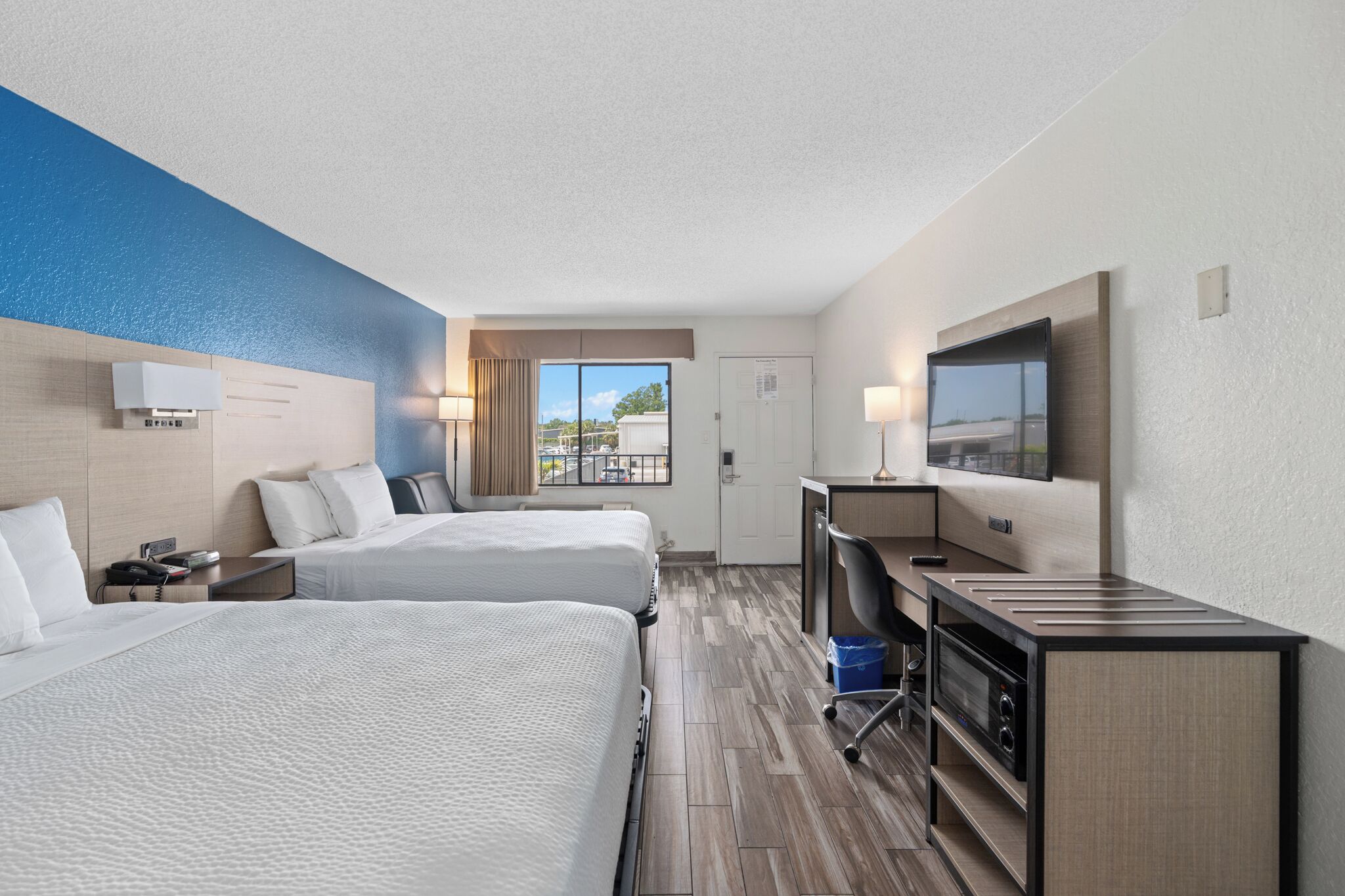 A hotel room with two beds, a desk with a flat-screen TV, a minibar, and a window with a view of the outside.