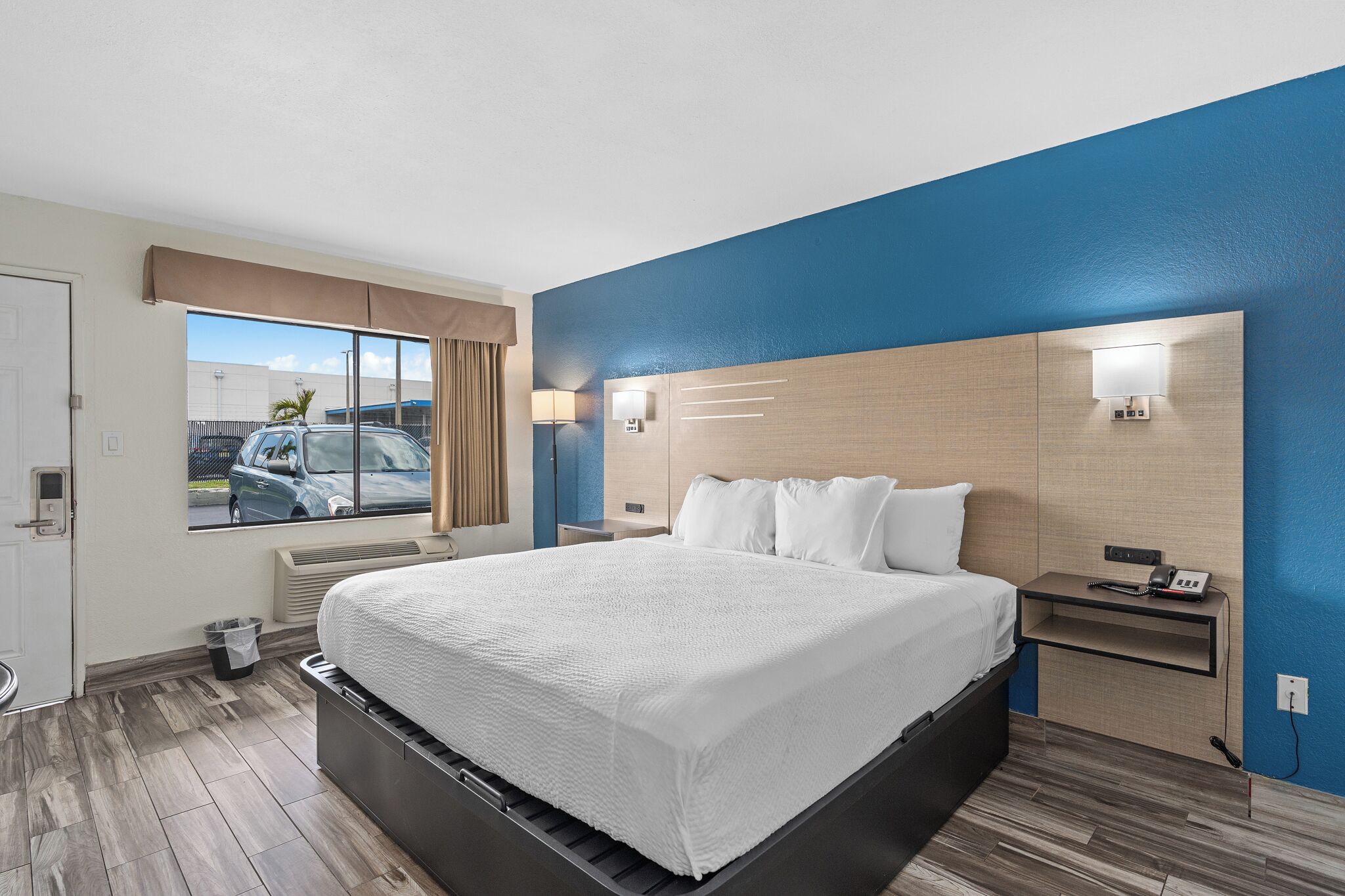 A modern hotel room with a large bed, a nightstand, a window with a view of the parking lot, and a blue accent wall.