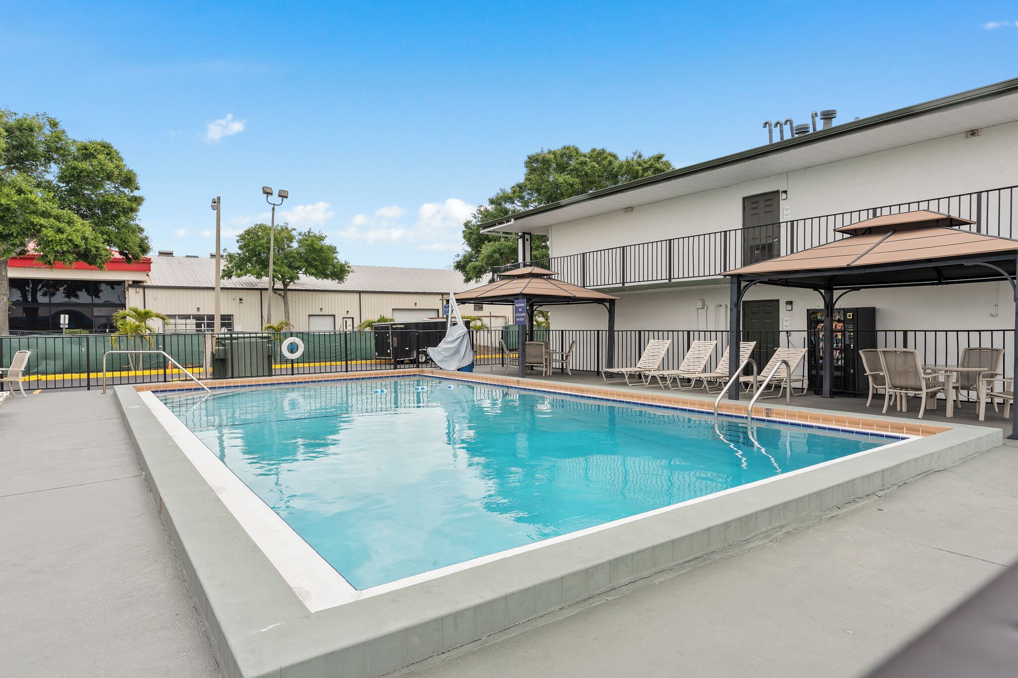 A rectangular pool with a clear blue water surface, surrounded by a concrete deck. There are lounge chairs and a covered seating area with a roof.