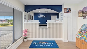 A reception area with a Baymont by Wyndham sign, a reception desk, a potted plant, a welcome mat, and a display of magazines. - Baymont by Wyndham Orlando Downtown (Orlando)