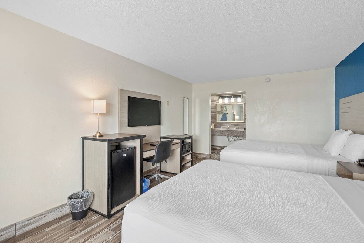 A hotel room with two beds, a desk, a chair, a TV, and a bathroom with a mirror and lights.