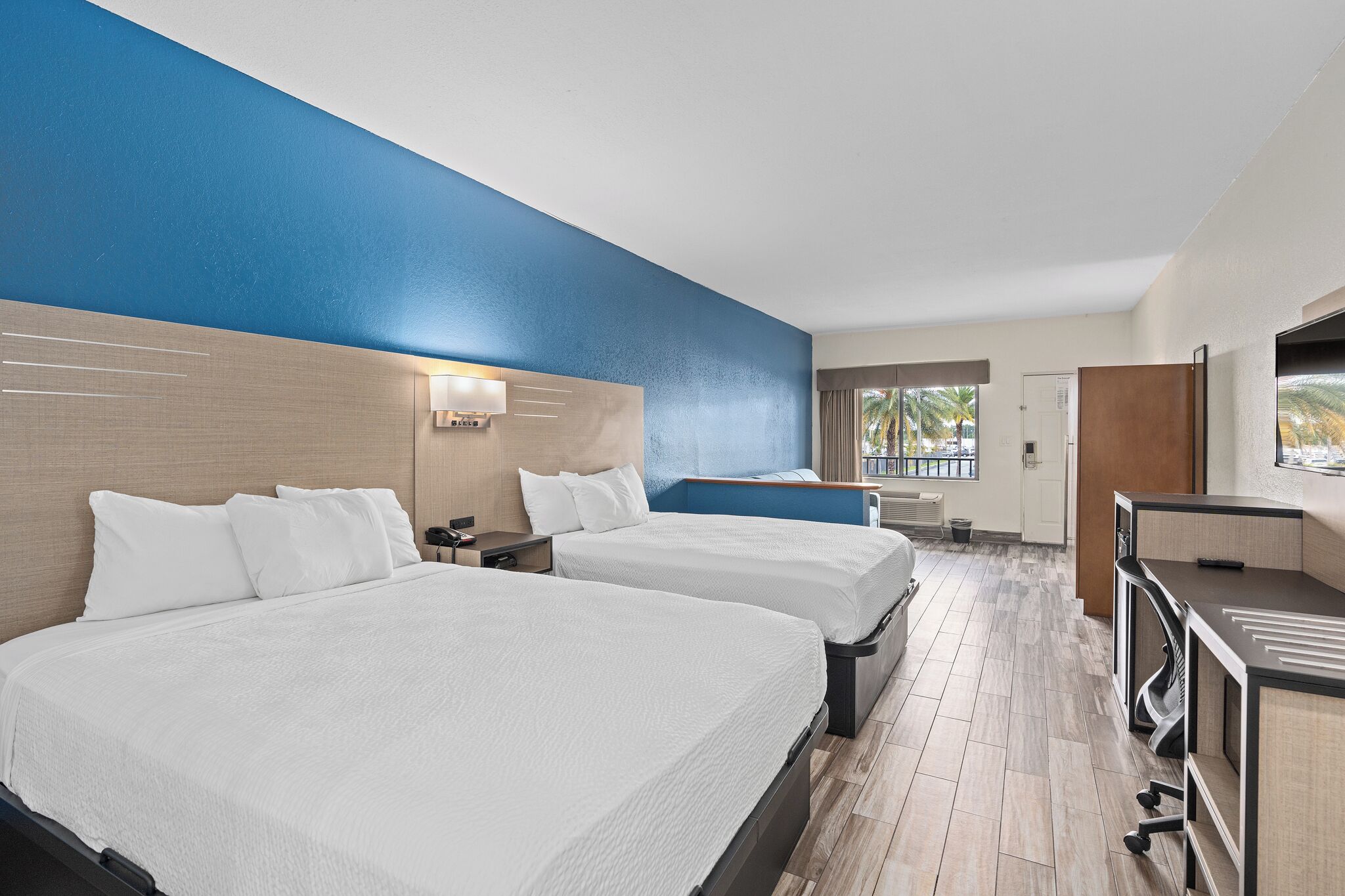 Habitación de hotel con dos camas, un escritorio y ventana con vista a palmeras.
