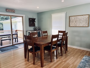 A dining area with a wooden table and chairs, a small kitchen area with a refrigerator and cabinets, and a view of the outdoors through a sliding glass door.