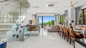 Uma sala de estar moderna com escada de vidro, área de jantar com mobiliário de madeira e vista para o oceano.