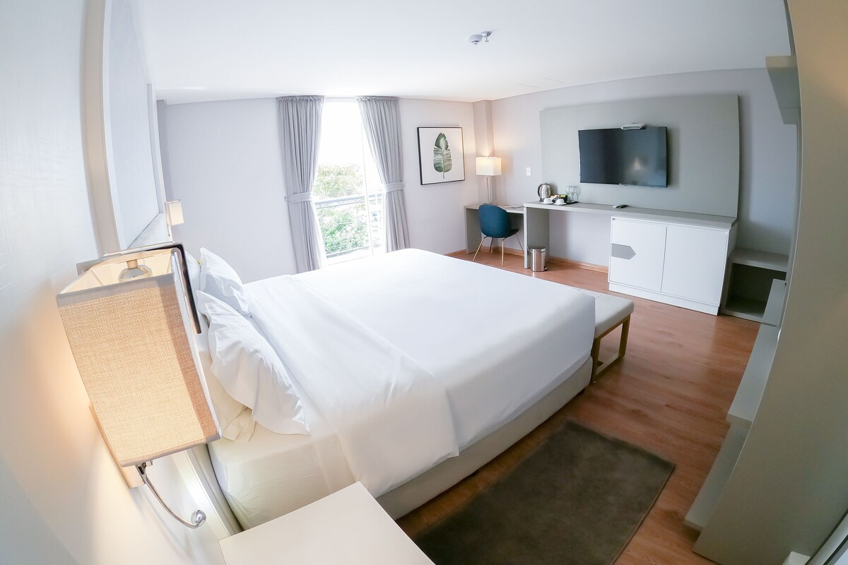 A hotel room with a large bed, a desk with a chair, a television, and a window with curtains.