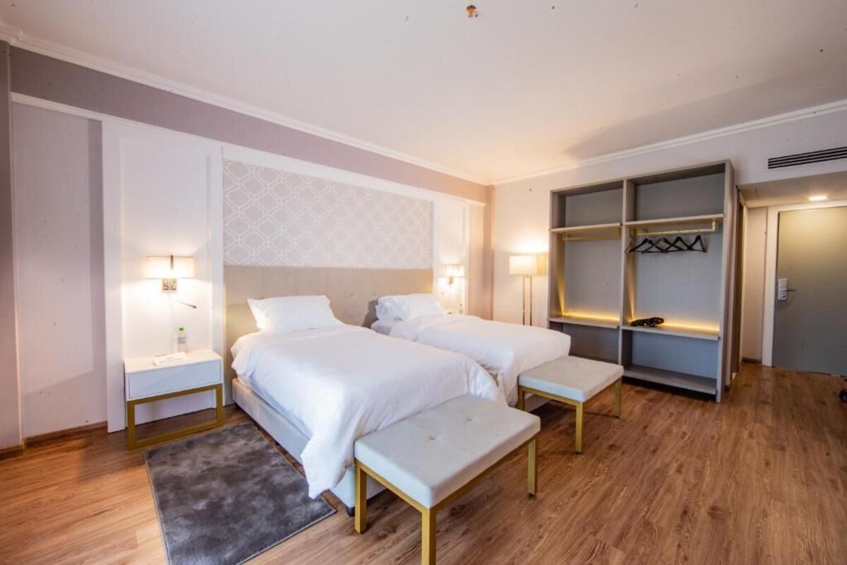 A hotel room with two beds, bedside tables, a bench, and a wardrobe.