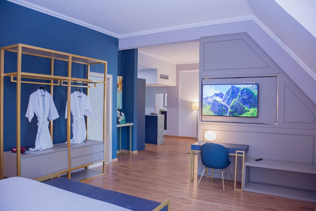 A modern hotel room with a wooden wardrobe, a flat-screen TV mounted on the wall, a desk with a chair, and a bed.