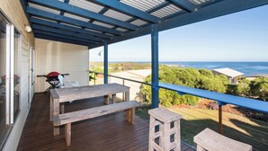 Um espaço ao ar livre coberto, com mesa e bancos de madeira, com vista panorâmica para o oceano e vegetação.
