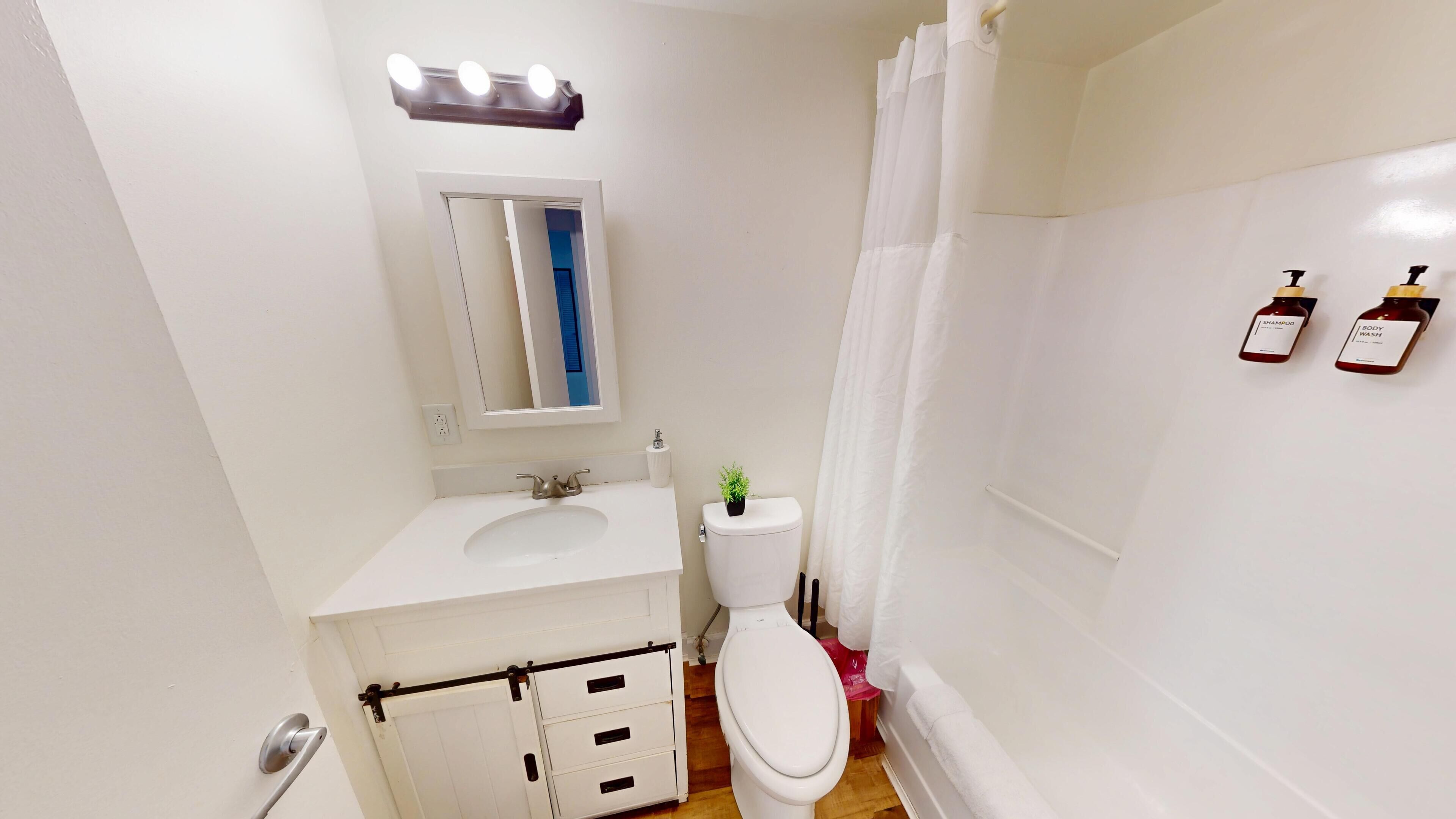 Un baño pequeño y limpio con lavamanos, inodoro y cortina de ducha blancos. Hay dos botellas de champú y gel de baño en la pared.