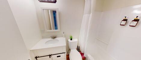 Un baño pequeño y limpio con lavamanos, inodoro y cortina de ducha blancos. Hay dos botellas de champú y gel de baño en la pared.