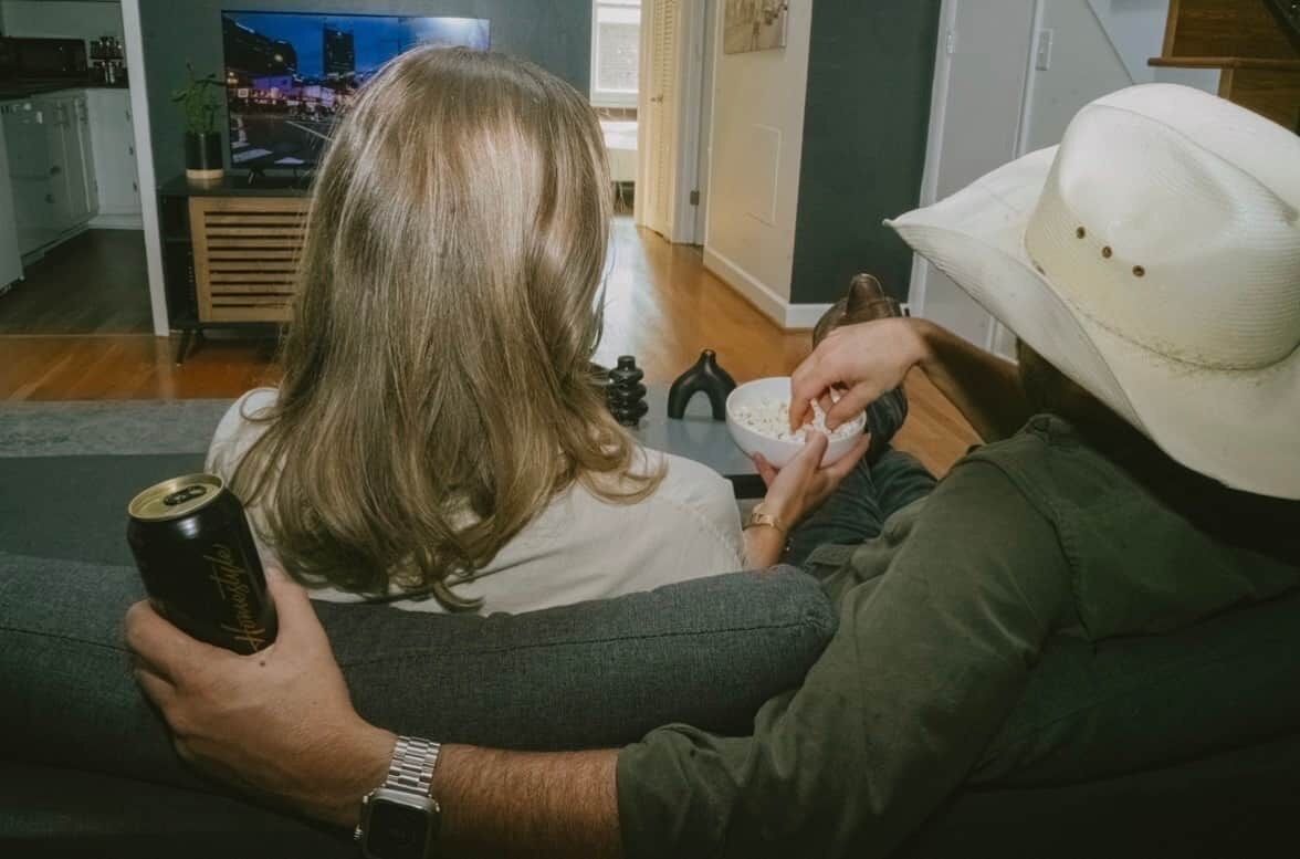 A person sitting on a couch, holding a can and a bowl, with another person in a cowboy hat nearby.
