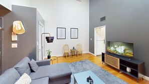 Un salon moderne avec un canapé gris, une table basse, un téléviseur posé sur un meuble en bois, deux tableaux encadrés accrochés au mur et une petite plante sur une table d’appoint.