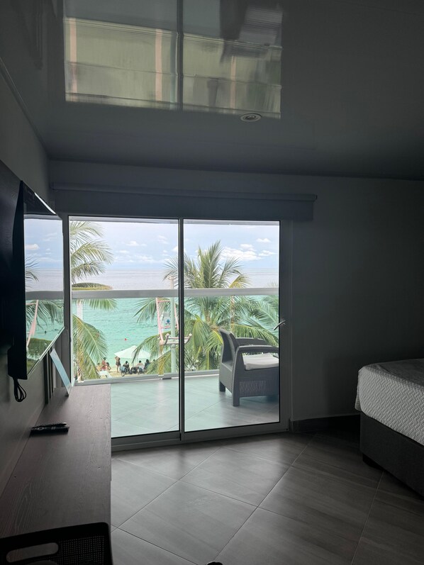 A room with a bed, a TV, and a large sliding glass door leading to a balcony with a view of the sea and palm trees. - Molino De Viento (San Andrés)