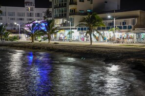 A beachfront hotel with a pool area, palm trees, and a well-lit walkway.