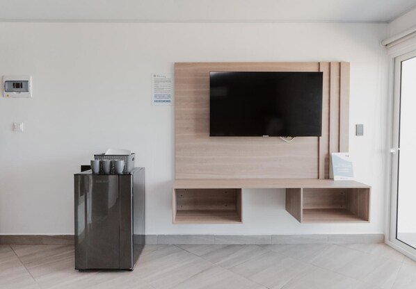 A modern living room with a flat-screen TV mounted on a wooden wall, a built-in kitchen unit with a refrigerator, and a glass door leading to a balcony. - Molino De Viento (San Andrés)