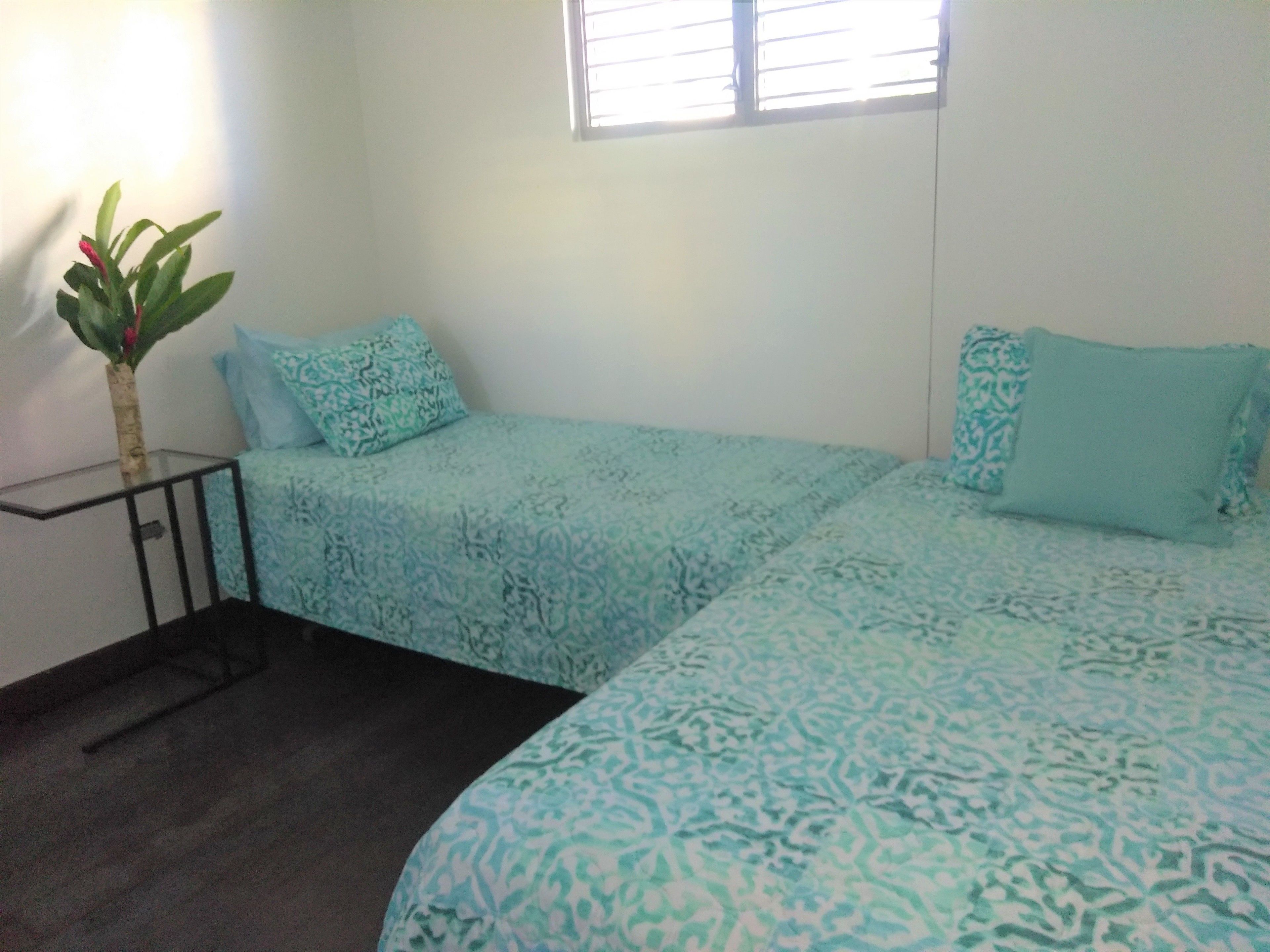 Een kleine, eenvoudige kamer met twee bedden, een nachtkastje, een bloempot en een raam met zonwering.