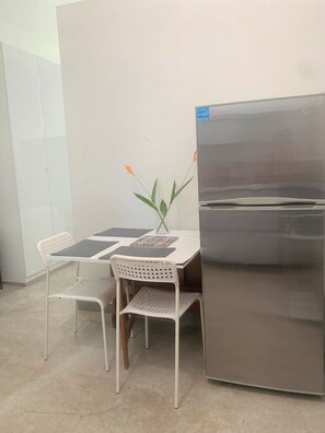 A small kitchenette with a stainless steel refrigerator, two white chairs, a table with black placemats, and a vase with green leaves and orange flowers.