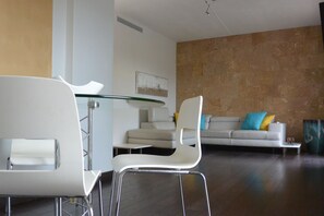 A modern living room with a glass-top dining table, white chairs, a sofa with colorful cushions, and a textured wall.