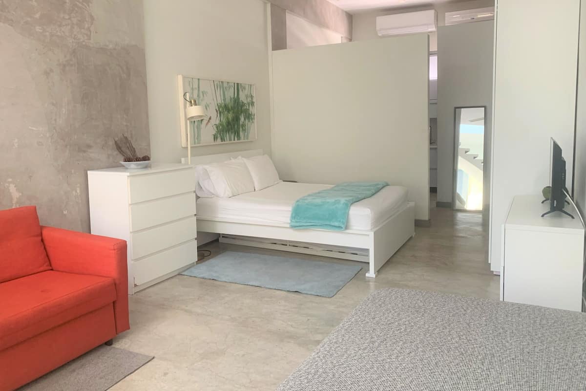 A modern bedroom with a bed, a red chair, a white dresser, and a painting on the wall.