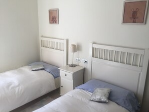 Two single beds with pillows and headboards, a bedside table with a lamp, and framed pictures adorn the walls.