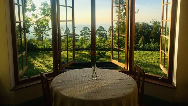 A room with a round table and a vase, large windows with wooden frames, and a view of greenery and trees. - Full board on an organic tea estate high in the hills - whole cottage with staff (Hildummulla)