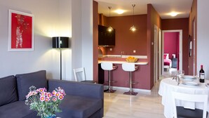 A modern living room with a grey sofa, a vase of flowers, and a red abstract painting. A kitchen with a white countertop and two bar stools is visible in the background. - Triana Terrace. 1-bedroom, roof-top terrace (Sevilla)