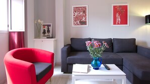 A living room with a red armchair, a grey sofa, a white coffee table with a vase of flowers, and two framed artworks on the wall. - Triana Terrace. 1-bedroom, roof-top terrace (Sevilla)