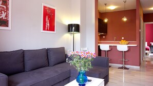 A modern living room with a grey sofa, a blue vase with flowers, a framed artwork, and a kitchen area with a red wall and white bar stools. - Triana Terrace. 1-bedroom, roof-top terrace (Sevilla)