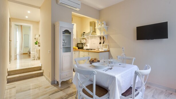 Un comedor con una mesa preparada para cuatro, sillas blancas, una zona de cocina con armarios y una estufa, y un televisor montado en la pared.