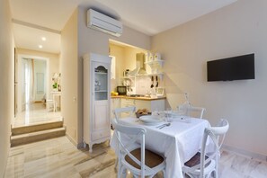 Un coin repas avec une table dressée pour quatre personnes, des chaises blanches, un espace cuisine avec des placards et une cuisinière, et un téléviseur fixé au mur.