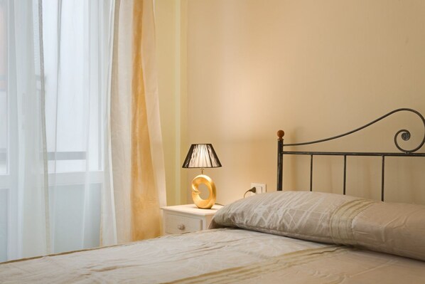 Un lit bien fait, recouvert d’une couvre-lit à motifs, une lampe de chevet au design original et une fenêtre ornée de rideaux légers.
