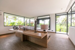 A modern room with a large wooden desk, a bed with white bedding, and large windows offering a view of greenery.