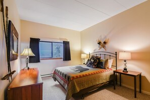 Une chambre à coucher comprenant un lit, deux tables de chevet, une fenêtre avec des rideaux, une lampe fixée au mur et un ventilateur de plafond.