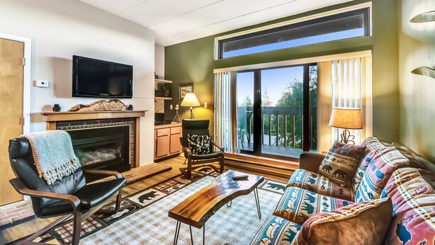 A living room with a fireplace, a television, a sofa, and a view of the outdoors. - Pinnacle A5 Condo | Pool, Hot Tub | Golf Nearby (Killington)